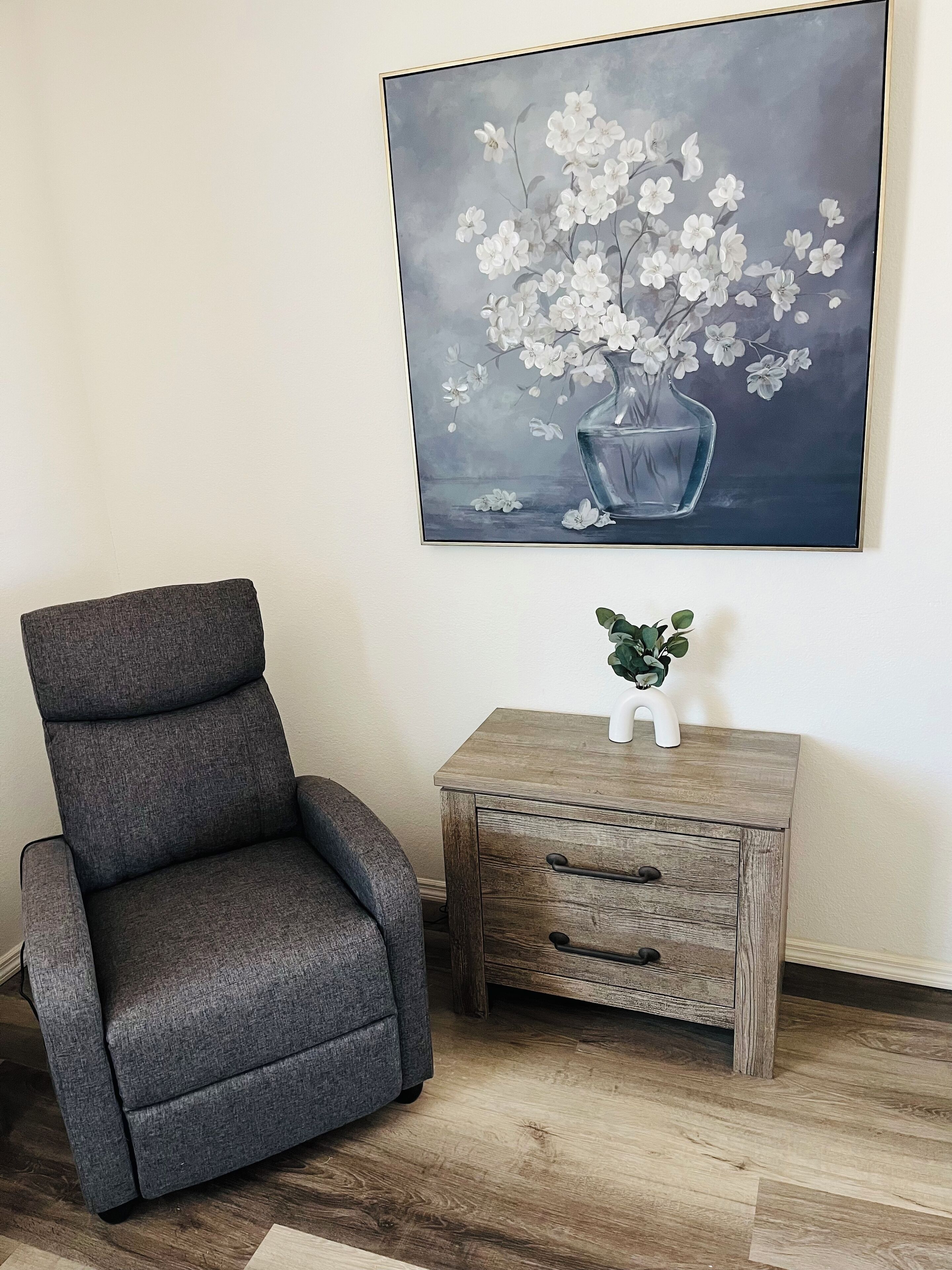 Un sillón gris y una mesita de noche de madera con un jarrón de flores blancas.