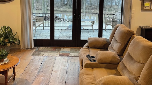 Una sala de estar acogedora con un sofá color beige, una mesa de centro de madera y un amplio ventanal corredero que da a un patio exterior con una silla blanca y una mesa.