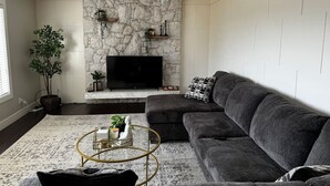 A living room with a stone wall, a flat-screen TV, a dark grey sofa, and a round coffee table. - Silver Oak Luxe Stay | King Bed | Retreat (Arrow Creek)