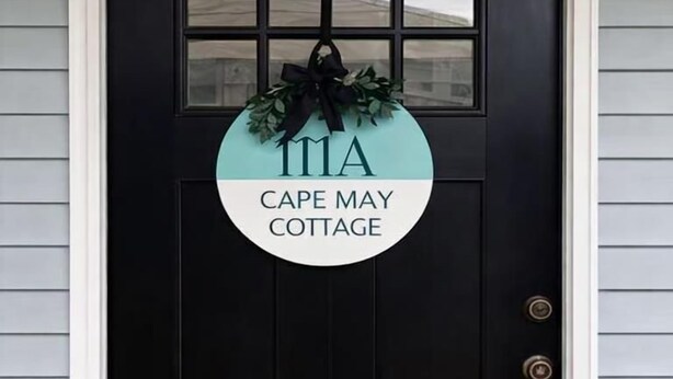 Una puerta negra con un cartel circular que dice "CAPE MAY COTTAGE" y una guirnalda encima.