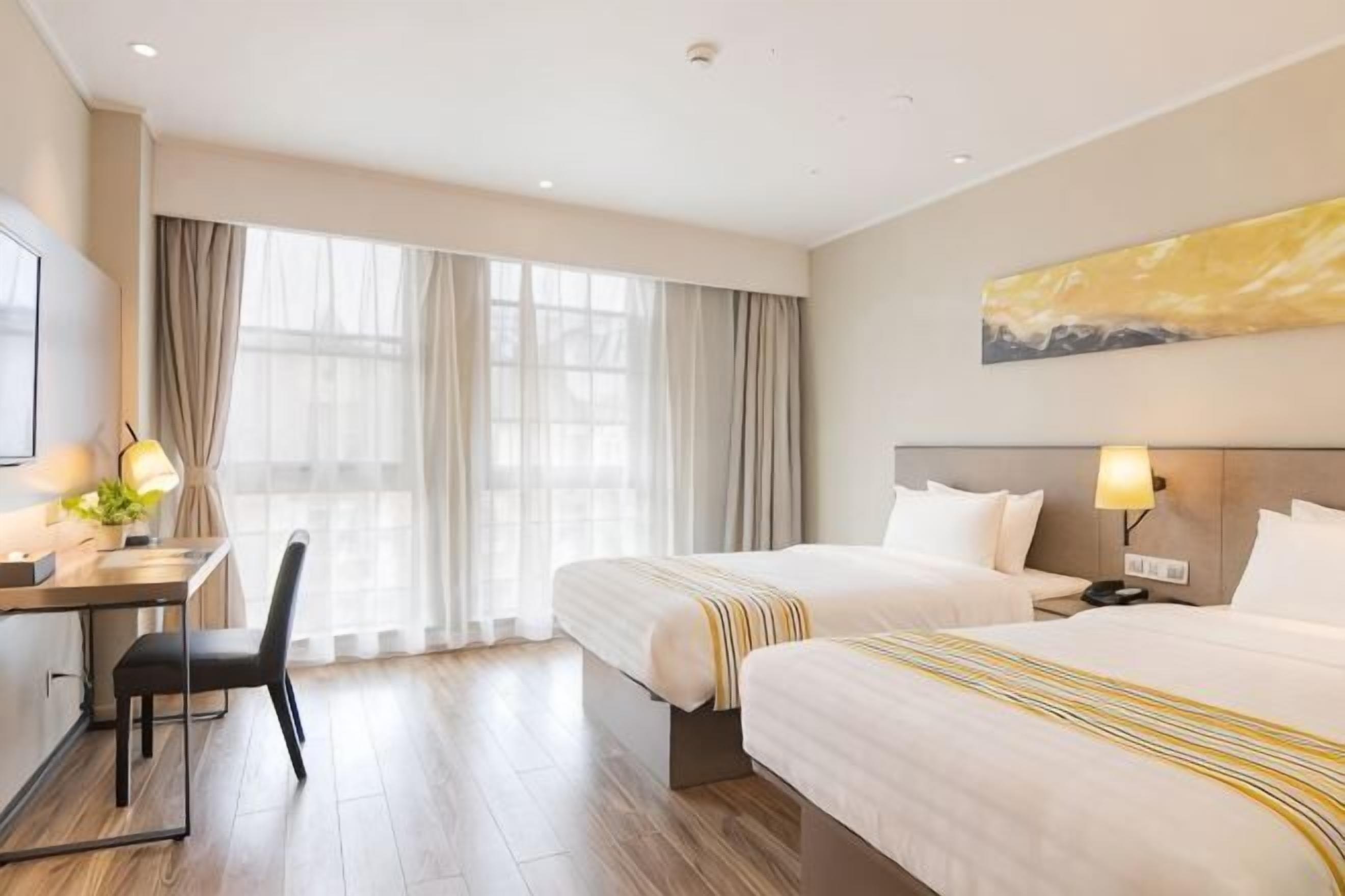 A hotel room with two beds, a desk, a chair, a TV, and a large window with curtains.