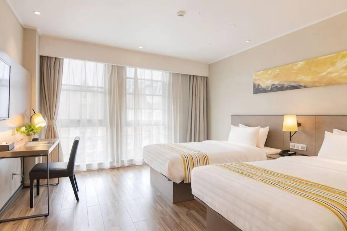A hotel room with two beds, a desk, a chair, a TV, and a large window with curtains.