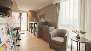 Une chambre d’hôtel moderne équipée d’un téléviseur à écran plat, d’un canapé, d’une petite table avec une plante, d’un lave-linge et d’une kitchenette.