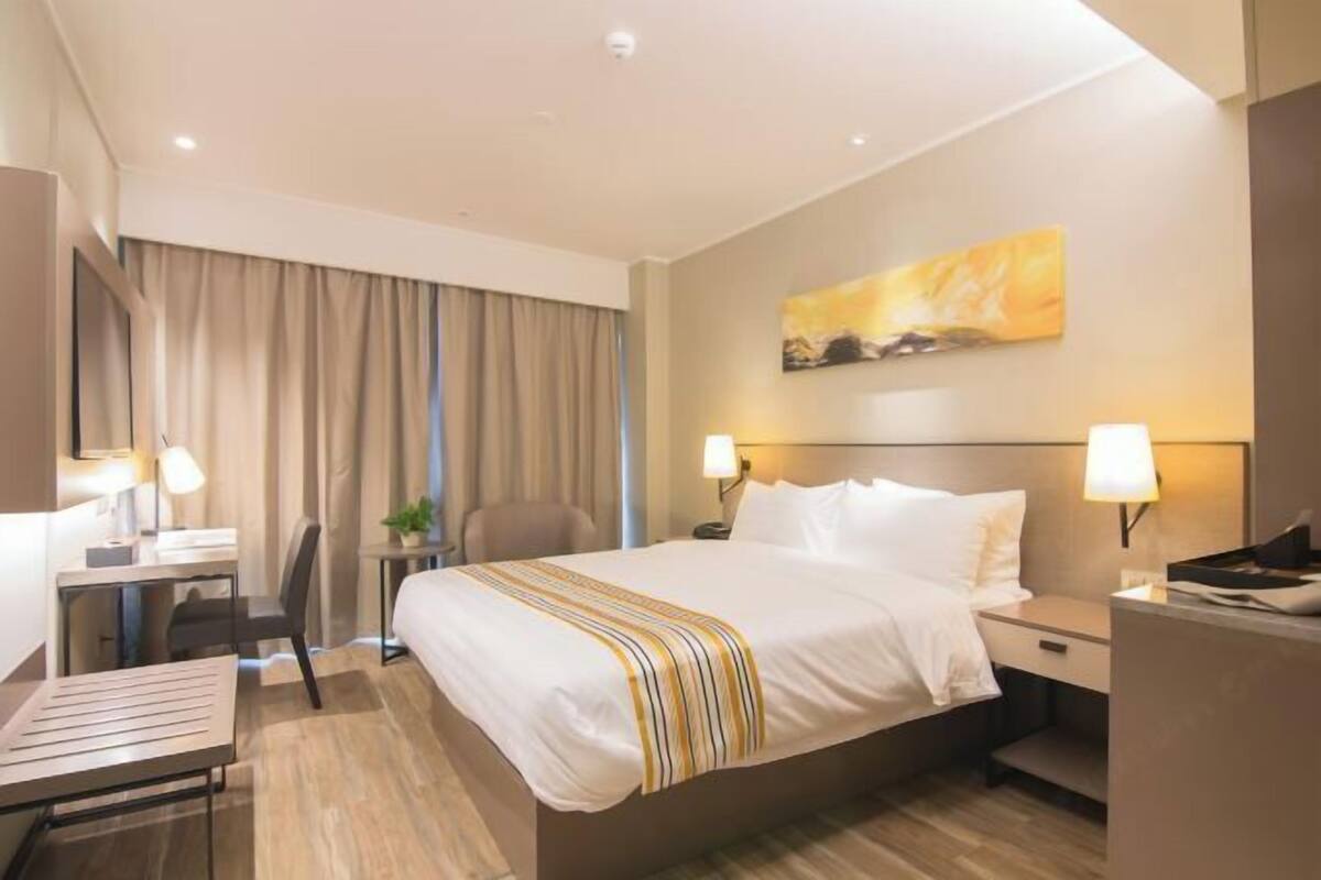 A hotel room with a bed, desk, chair, and a painting on the wall.