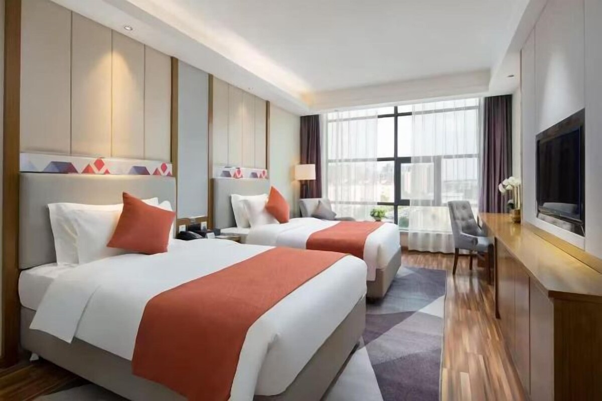 A hotel room with two beds, a TV, a desk, and a large window.