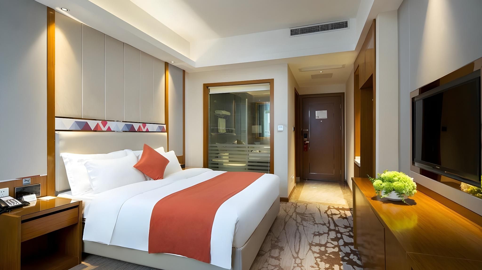 A hotel room with a bed, a desk, a TV, and a bathroom visible through a glass door.