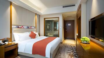 A hotel room with a bed, a desk, a TV, and a bathroom visible through a glass door.