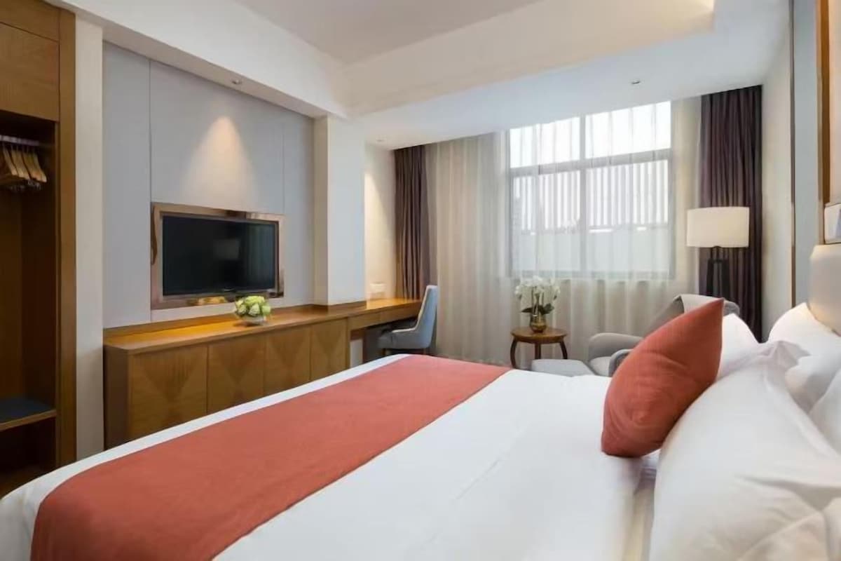 A hotel room with a bed, a desk with a television, a chair, a small table with a vase, and a window with curtains.