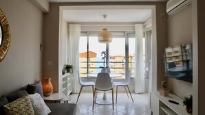 Una sala de estar moderna con un sofá, una mesa de comedor con sillas, un televisor montado en la pared y un amplio ventanal con vistas a edificios.