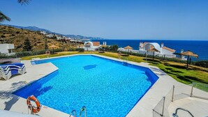 A clear blue swimming pool with a lifebuoy, a ladder, and a lifeguard stand. In the background, there are white buildings with red roofs and a view of the sea.