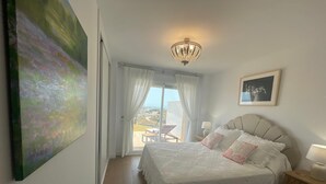 A bedroom with a large bed, a chandelier, a painting, and a view through the window.