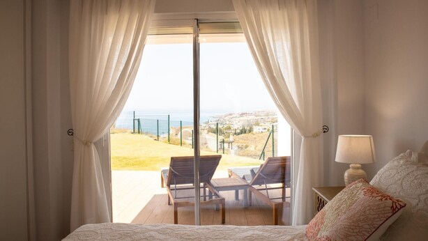 A bedroom with a bed, a nightstand with a lamp, and a view of a balcony with chairs and a table.