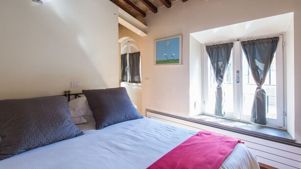 A bedroom with a bed, a window with curtains, a painting on the wall, and a ceiling with exposed beams.