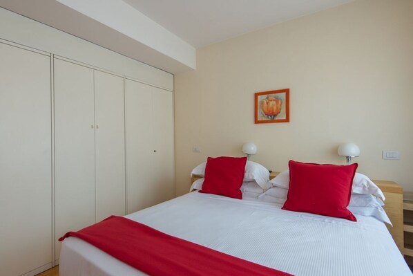 Un dormitorio con una cama, dos lámparas de noche, una colcha roja, un almohadón rojo, un cabecero blanco y un cuadro en la pared.