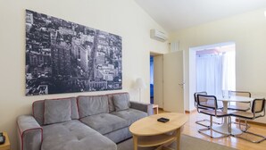 A living room with a gray sofa, a round coffee table, a dining table with chairs, a large black-and-white cityscape painting, and a door leading to another room.