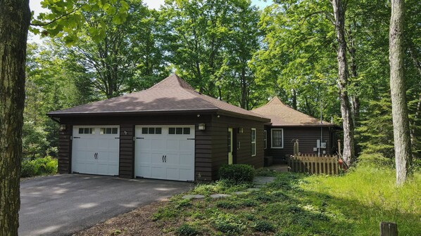 Eine zweistöckige Holzhütte mit Garage und kleiner Veranda, umgeben von dichtem Wald.