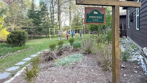 A cabin named Pinecone with a wooden sign, surrounded by a garden with stepping stones, a volleyball net, and trees. - Updated cabin sleeps 8. Pet friendly, and has private neighborhood beach. (Charlevoix)