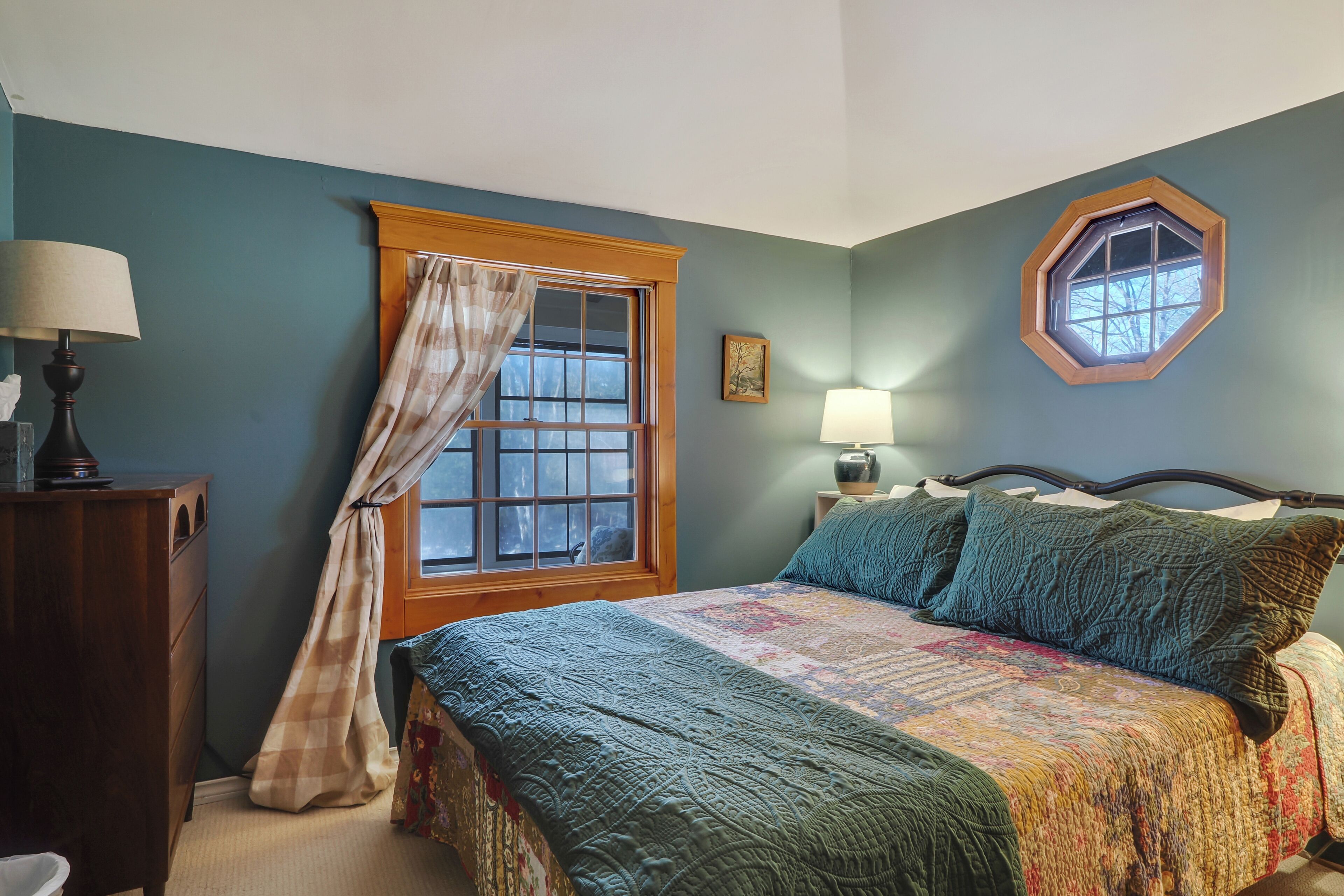 Un dormitorio con cama, una ventana con cortinas, una mesita de noche con lámpara y un aparador.