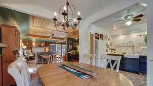 A dining area with a wooden table, white chairs, a chandelier, and a view into the kitchen area with blue cabinets and a ceiling fan. - Updated cabin sleeps 8. Pet friendly, and has private neighborhood beach. (Charlevoix)