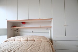 Een slaapkamer met een bed, een nachtkastje, een rode lamp en een witte garderobe met lades.