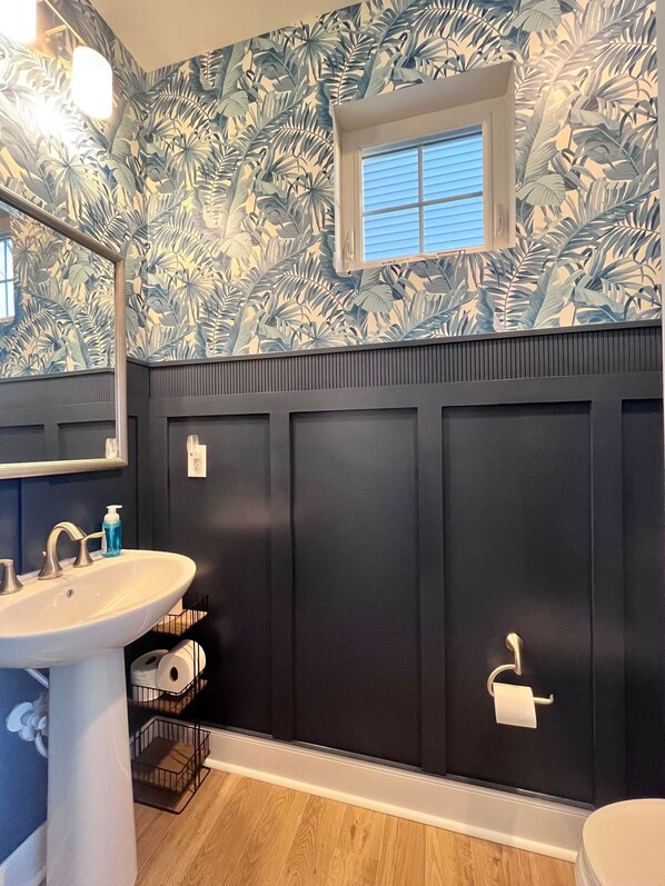 A bathroom with a white sink, a mirror, and a toilet. The walls are dark blue with a tropical leaf pattern.