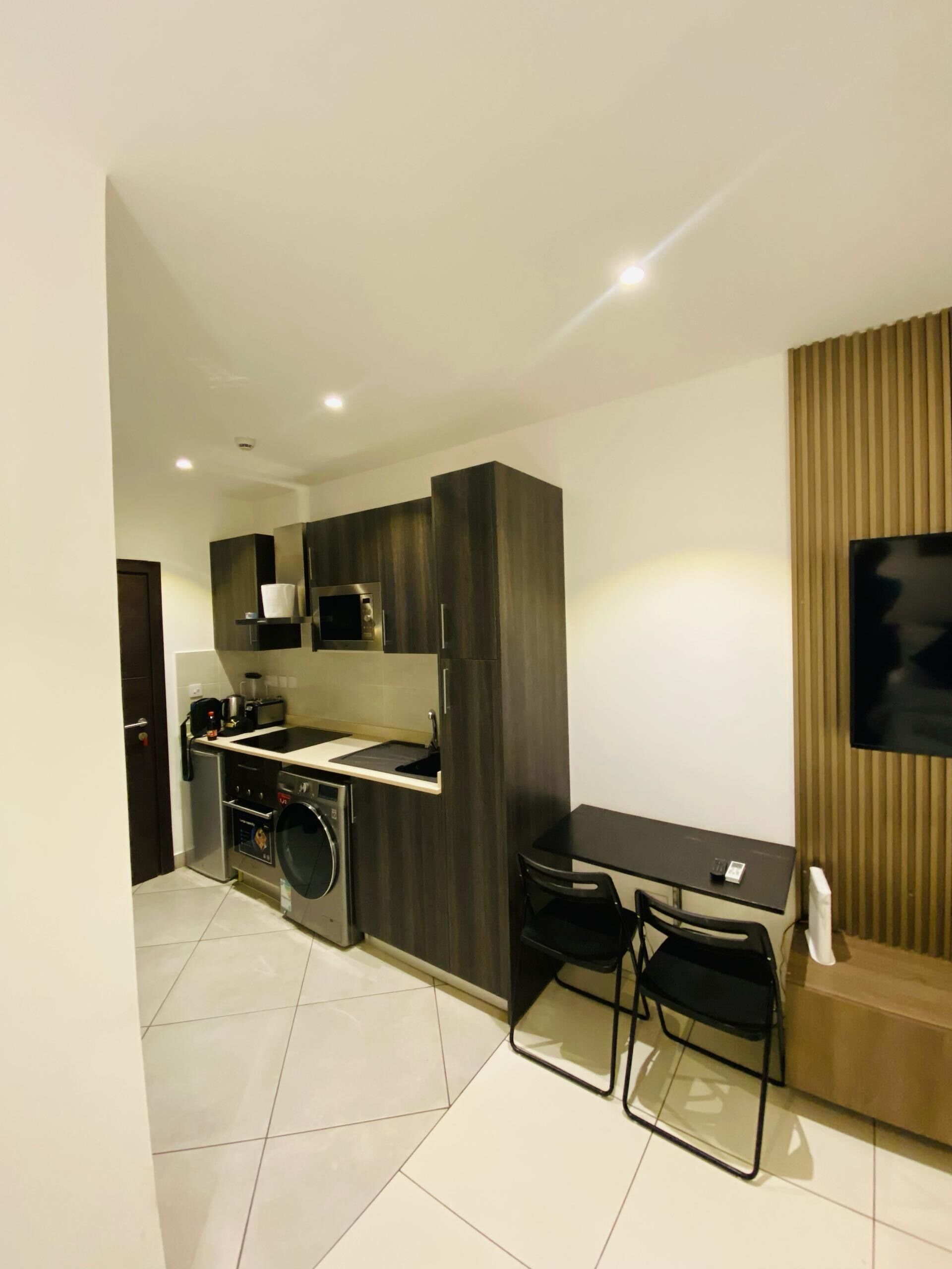 A compact kitchen with modern appliances, a dining area with a table and chairs, and a large TV mounted on the wall.