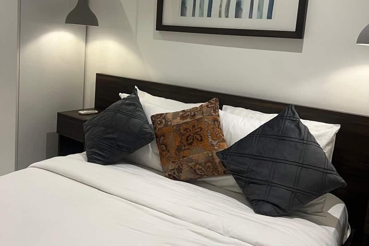 A neatly made bed with white bedding and multiple pillows, a framed artwork above it, and a bedside lamp on a nightstand.