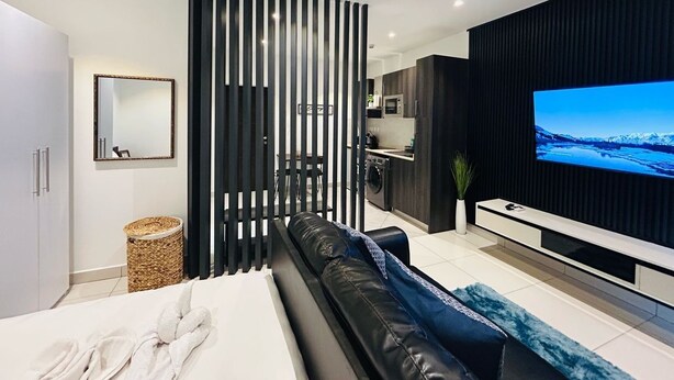 A modern bedroom with a bed, a TV, a sofa, and a kitchen area in the background. - EBS LEGEND ONE BEDROOM DUPLUX embassy (Accra)