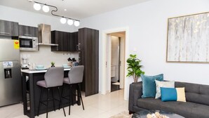 A modern kitchen with dark cabinets, a microwave, and a dining area with two chairs. There's a gray sofa with blue and beige cushions in the living area, and a plant on the floor. - EBS LEGEND ONE BEDROOM DUPLUX embassy (Accra)