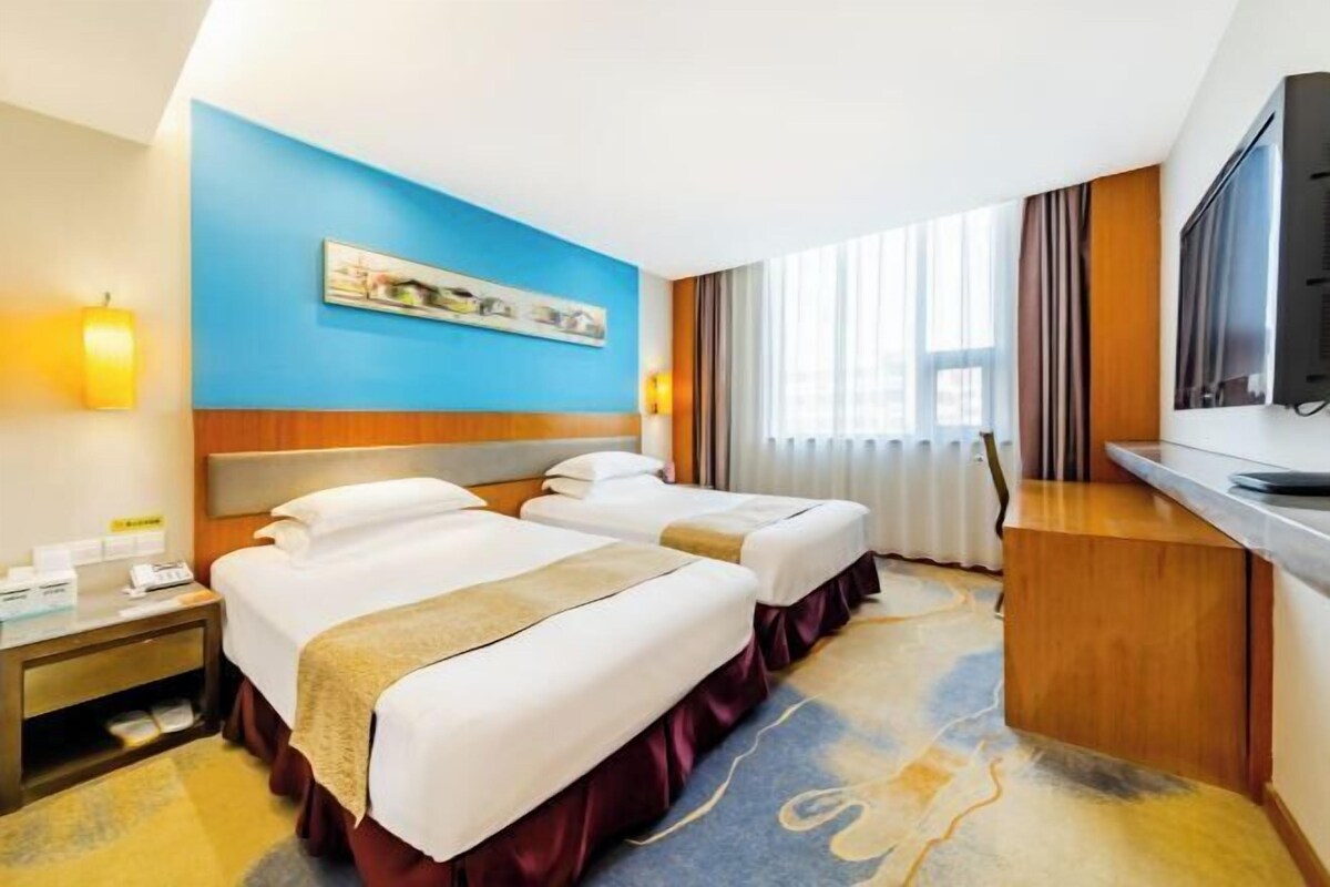 A hotel room with two beds, a desk, a television, and a large window with curtains.