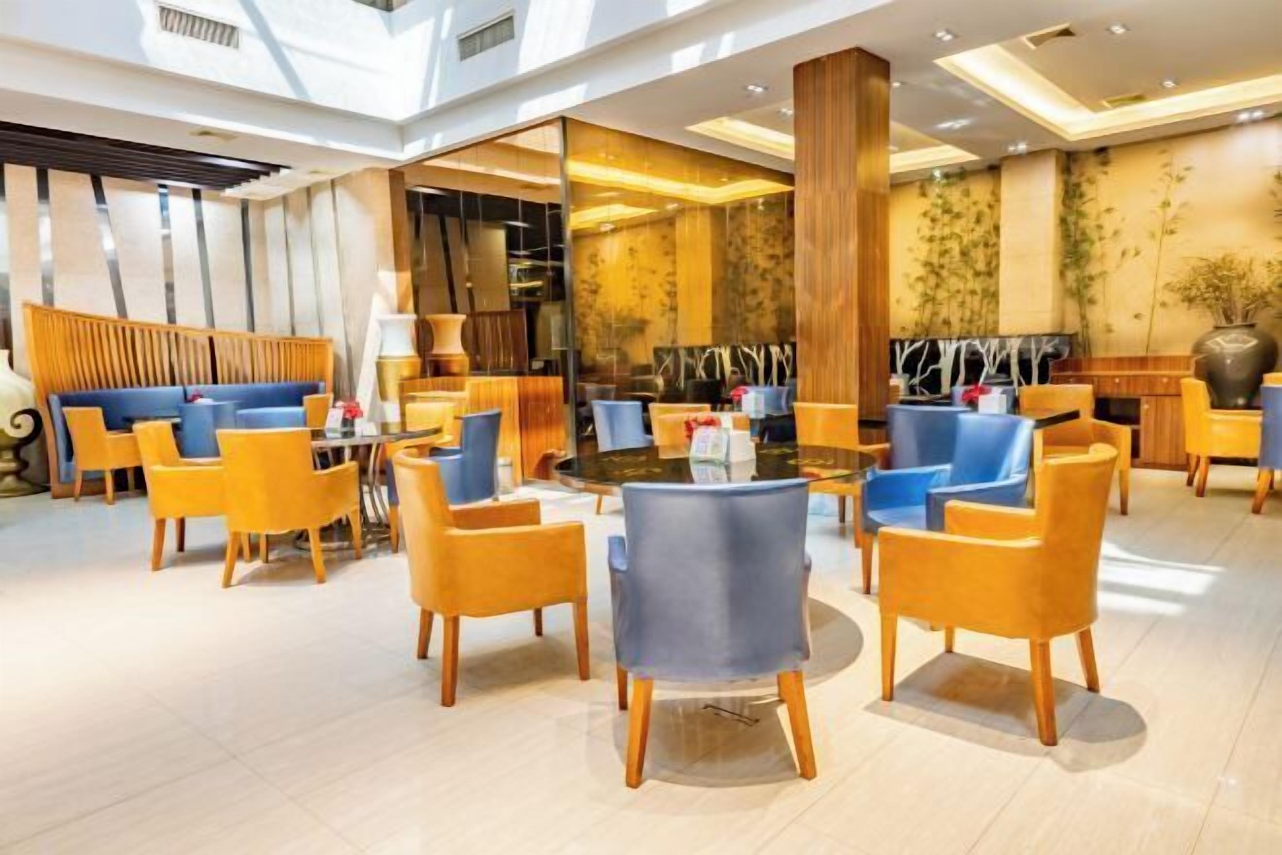 A modern hotel lobby with a central round table surrounded by yellow and blue chairs, wooden accents, and a large vase of flowers.