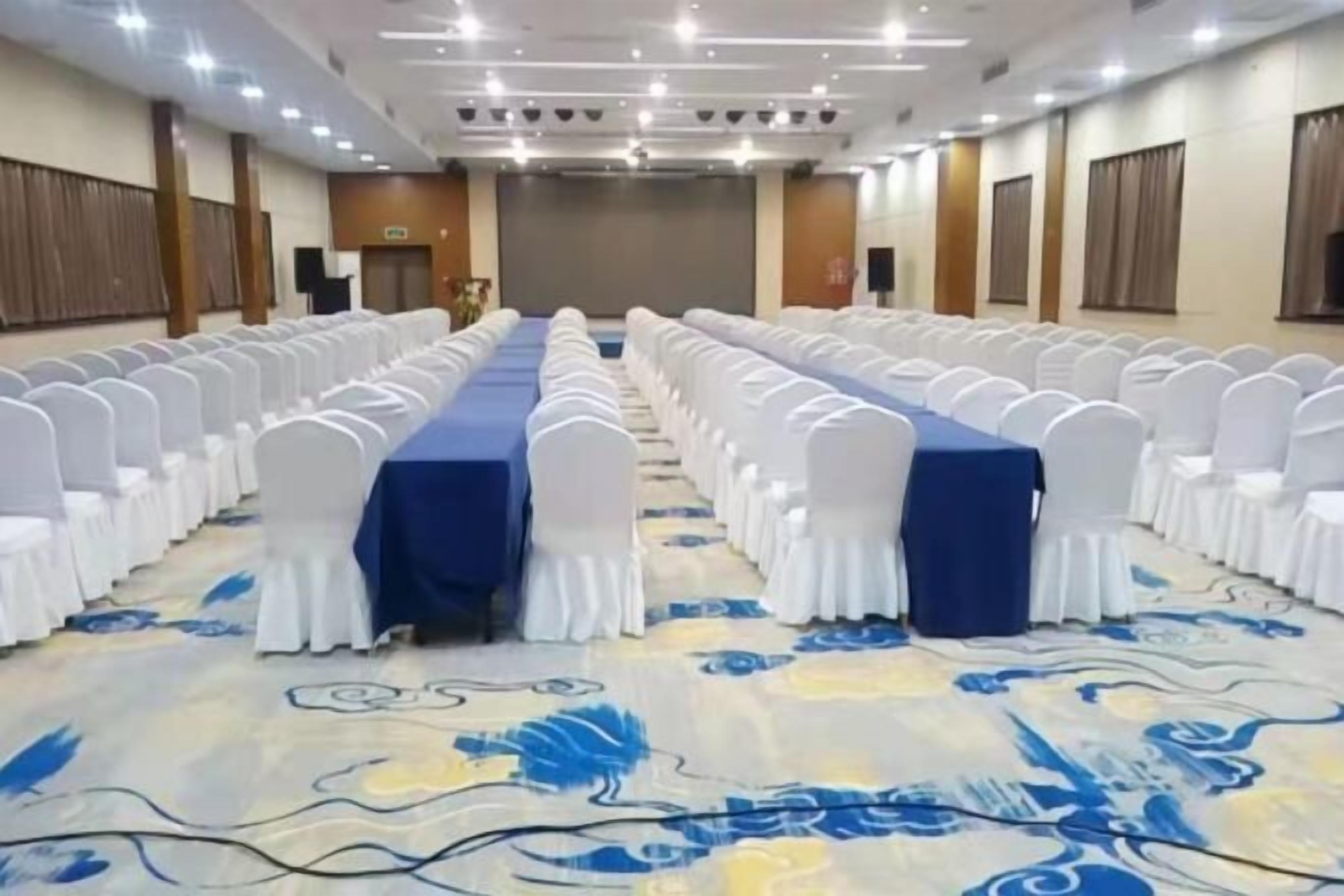 A conference room with rows of white chairs, a blue table, and a patterned carpet.