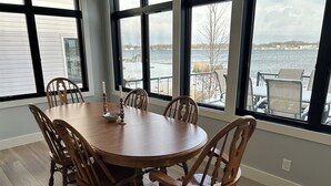 Un coin repas avec une table ronde en bois et six chaises, de grandes fenêtres offrant une vue sur un plan d’eau et un quai.