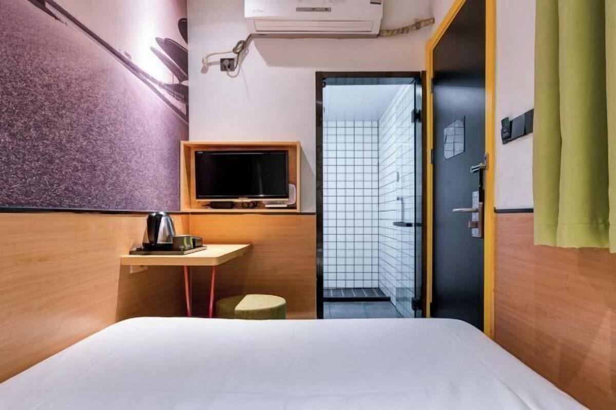 A hotel room with a bed, a small desk with a kettle, a television, and a door leading to a tiled area.