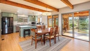 A modern kitchen with wooden beams, a dining area with a large table and chairs, and a sliding glass door leading to a patio. - Sundance Ranch Retreat | Hot Tub • Cascade Views (Deschutes County)
