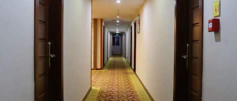 A hotel hallway with carpeted floors, wooden doors, and a fire alarm on the wall.