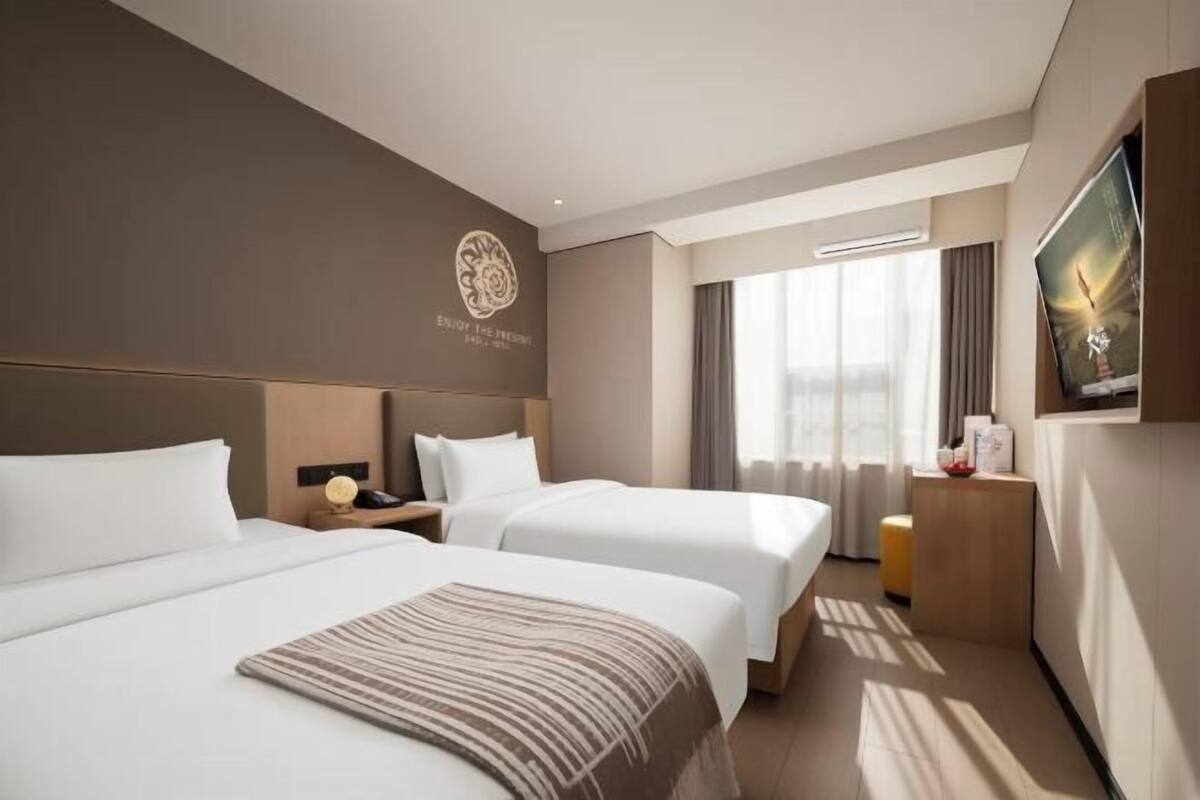 Ein Hotelzimmer mit zwei Betten, einem großen Fenster mit Vorhängen, einem Flachbildfernseher und einem an der Wand befestigten Logo.
