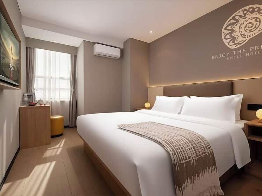 A hotel room with a large bed, a bedside table with a lamp, a wall-mounted air conditioner, and a framed picture on the wall.