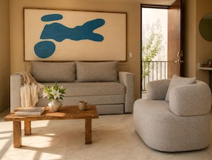 Un salon comprenant un canapé, un fauteuil rond, une table basse avec un livre et un vase, ainsi qu’un grand tableau abstrait.