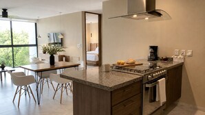 Moderni keittiö, jossa on keskeinen saareke, ruokailutila tuoleineen ja näkymä makuuhuoneeseen avonaisen oven kautta.