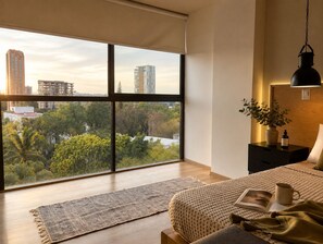 Une chambre avec une grande fenêtre, un lit, une table de chevet avec une lampe, et une vue sur le paysage urbain.