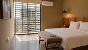 A bedroom with a large bed, a bedside table with a lamp, a window with blinds, and an air conditioning unit.