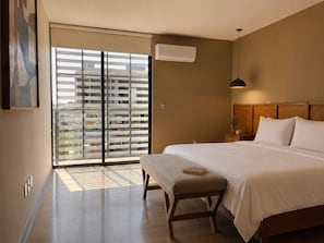 A bedroom with a large bed, a bedside table with a lamp, a window with blinds, and an air conditioning unit.
