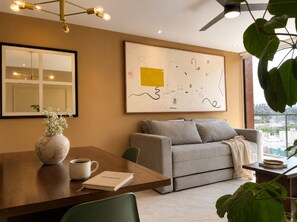 A modern living room with a sofa, a coffee table, a vase with flowers, a framed abstract painting, and a large window with a city view. - High Life suite / Daily Cleaning (guadalajara)