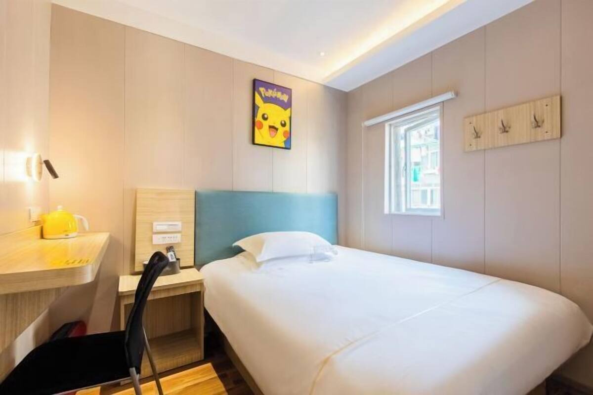 Habitació d'hotel amb un llit, una taula de treball amb una cadira, una taula petita amb un objecte groc i una finestra amb vistes.