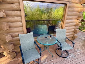 A log cabin with a glass sliding door, two chairs, a small round table, and a view of a forested mountain range.