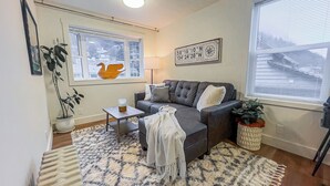 Ein Wohnzimmer mit grauem Sofa, einem kleinen Tisch, einem Couchtisch, einem gemusterten Teppich, einer Topfpflanze und einem Fenster mit Jalousien.