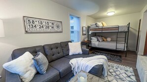Ein kompakter Wohnbereich mit einer Couch, einem Etagenbett, einem kleinen Tisch und einem an der Wand befestigten Kompass.