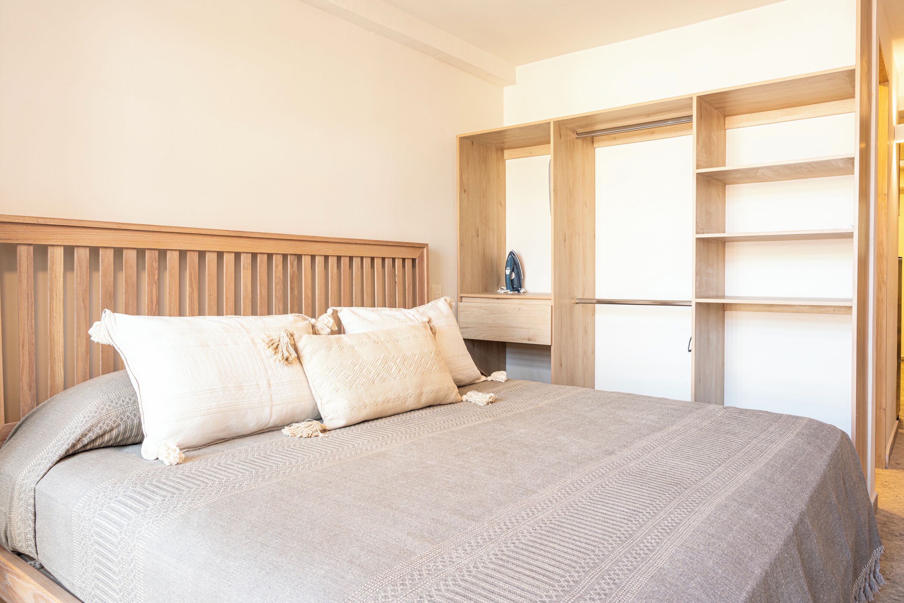 A neatly made bed with a headboard, a wooden wardrobe with open doors, and a small shelf with an iron.