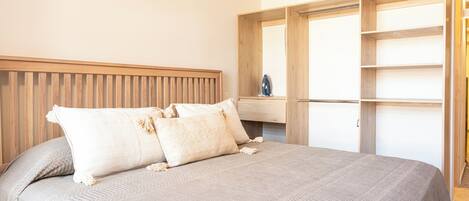 A neatly made bed with a headboard, a wooden wardrobe with open doors, and a small shelf with an iron.